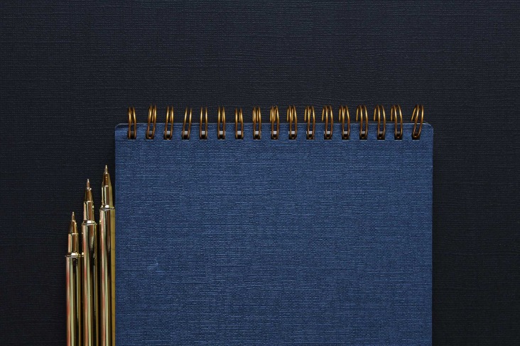 3 gold pens beside a wire-bound notebook on top of a cloth textured surface